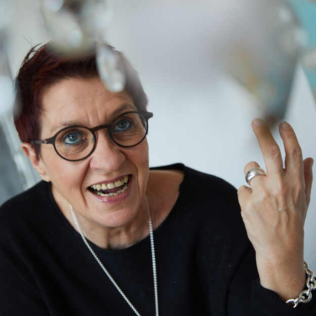 Lachende Frau mit kurzen, roten Haaren und Brille, die mit einer Hand gestikuliert, vor einem bunten Hintergrund.