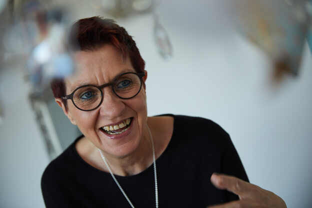 Lächelnde Frau mit Brille in schwarzem Oberteil, gestikuliert in einem hellen, modernen Raum.