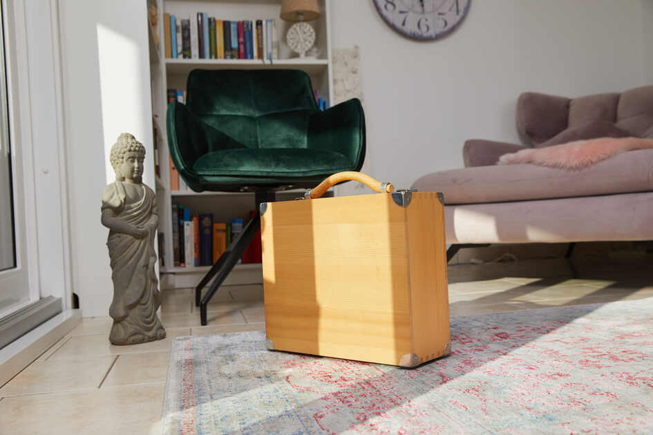 Holzkoffer steht auf einem Teppich neben einer Buddha-Statue und einem grünen Stuhl in einem hellen Raum.
