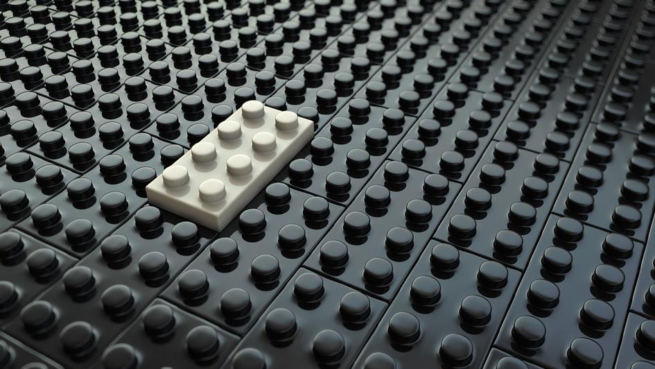 Ein einzelner grauer LEGO-Stein liegt auf einer Fläche aus schwarzen LEGO-Steinen.