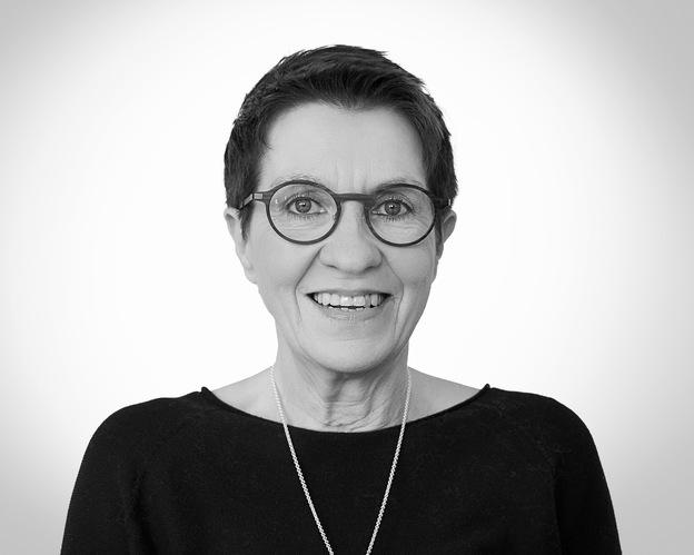 Porträt einer lächelnden Frau mit Kurzhaarschnitt, Brille und schwarzem Oberteil vor neutralem Hintergrund.